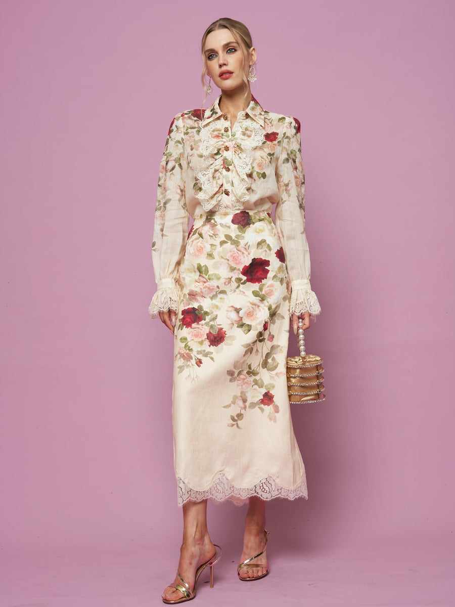 A woman stands against a pink background wearing the Social Queen Rose Print Ruffle Lace Blouse, paired with pink high heels and holding a small round handbag.