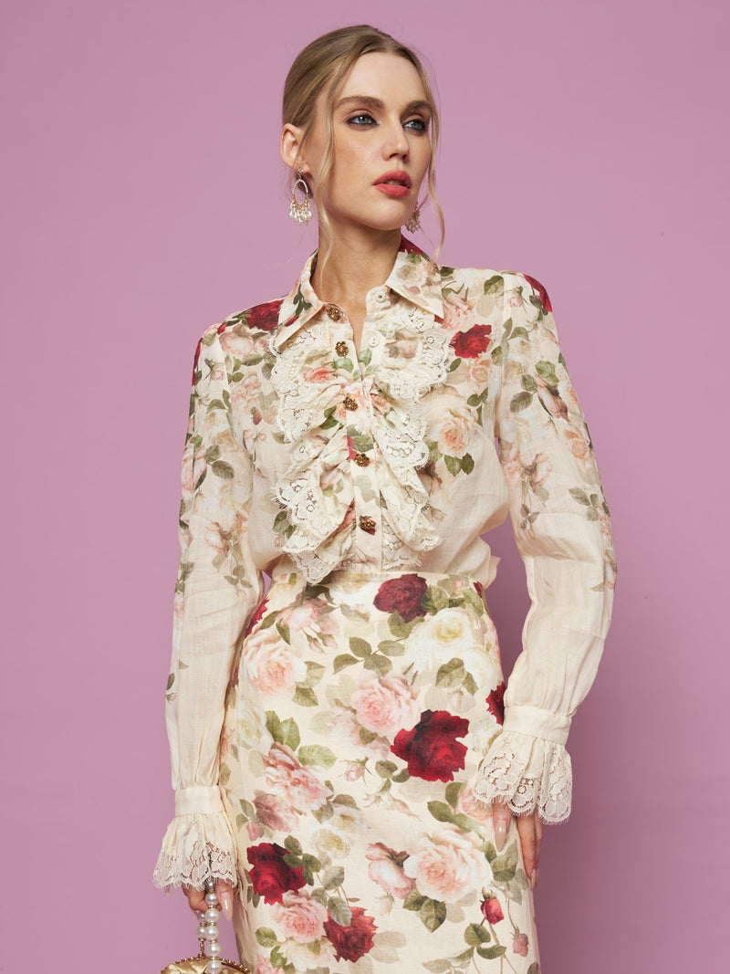 A woman stands against a pink background wearing the Social Queen Rose Print Ruffle Lace Blouse, featuring lace details and an oil-painting rose print.