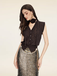 A woman in the Social Queen Rose-Tie Neck Knit Cardigan and a sequined silver skirt with chain belt, large earrings, and a floral neck accessory looks downward against a plain background.