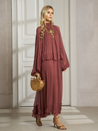 A woman stands indoors in the Social Queen Ruched High-Neck Ruffled Midi Dress, accessorized with a wooden handbag, heeled sandals, and a statement necklace.