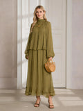 A woman stands indoors in the Social Queen Ruched High-Neck Ruffled Midi Dress—an olive green, pleated design with lantern sleeves—paired with a woven handbag and heeled sandals.