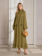 A woman stands indoors in the Social Queen Ruched High-Neck Ruffled Midi Dress—an olive green, pleated design with lantern sleeves—paired with a woven handbag and heeled sandals.