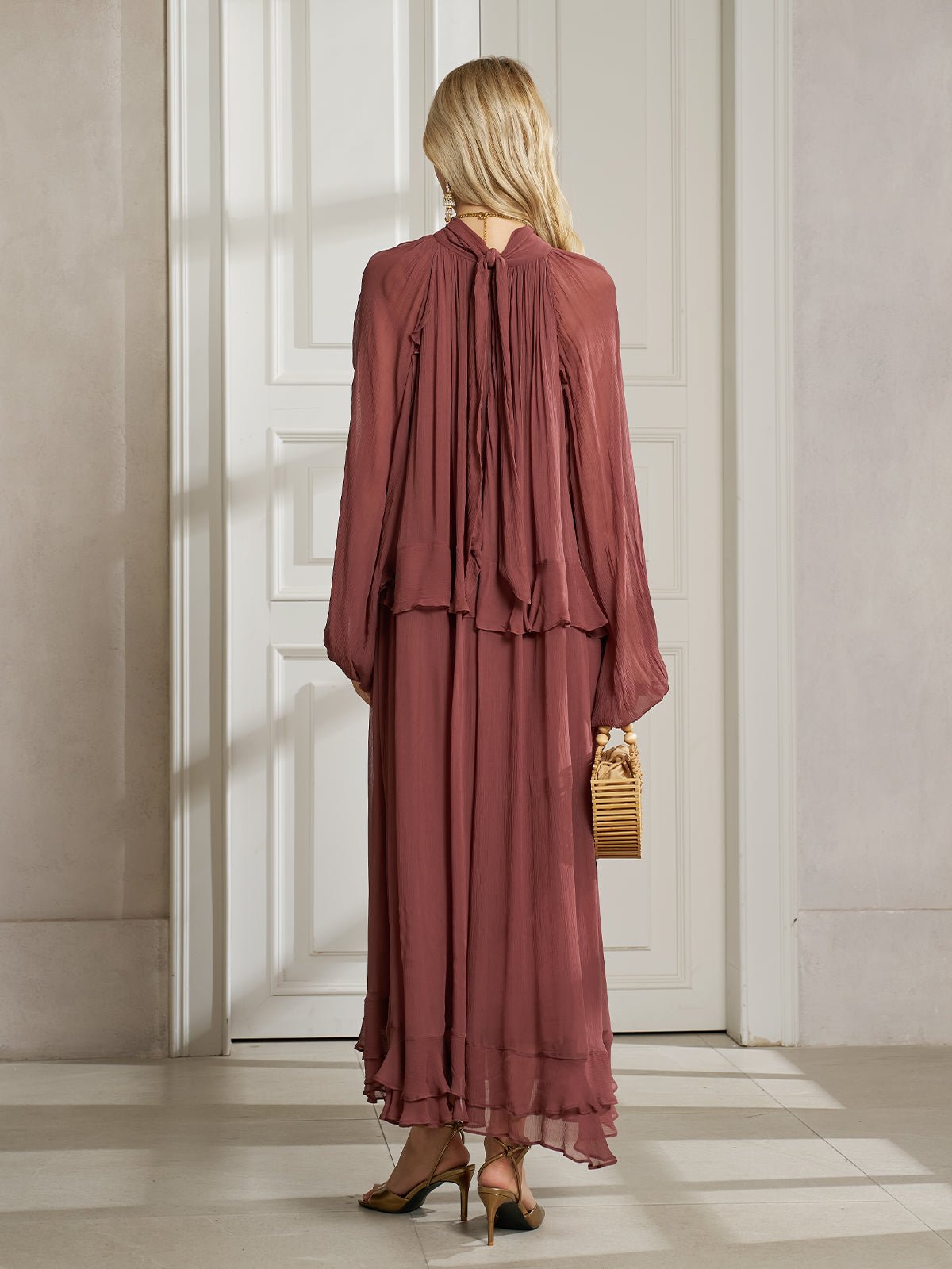 A woman stands indoors with her back to the camera, wearing the Social Queen Ruched High-Neck Ruffled Midi Dress in mauve and holding a small wooden handbag.