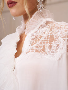 Close-up of a woman wearing the Social Queen Ruffled Lace Blouse with Asymmetric Hem, featuring organza ruffles at the collar and pearl earrings; only part of her face and upper body are visible.