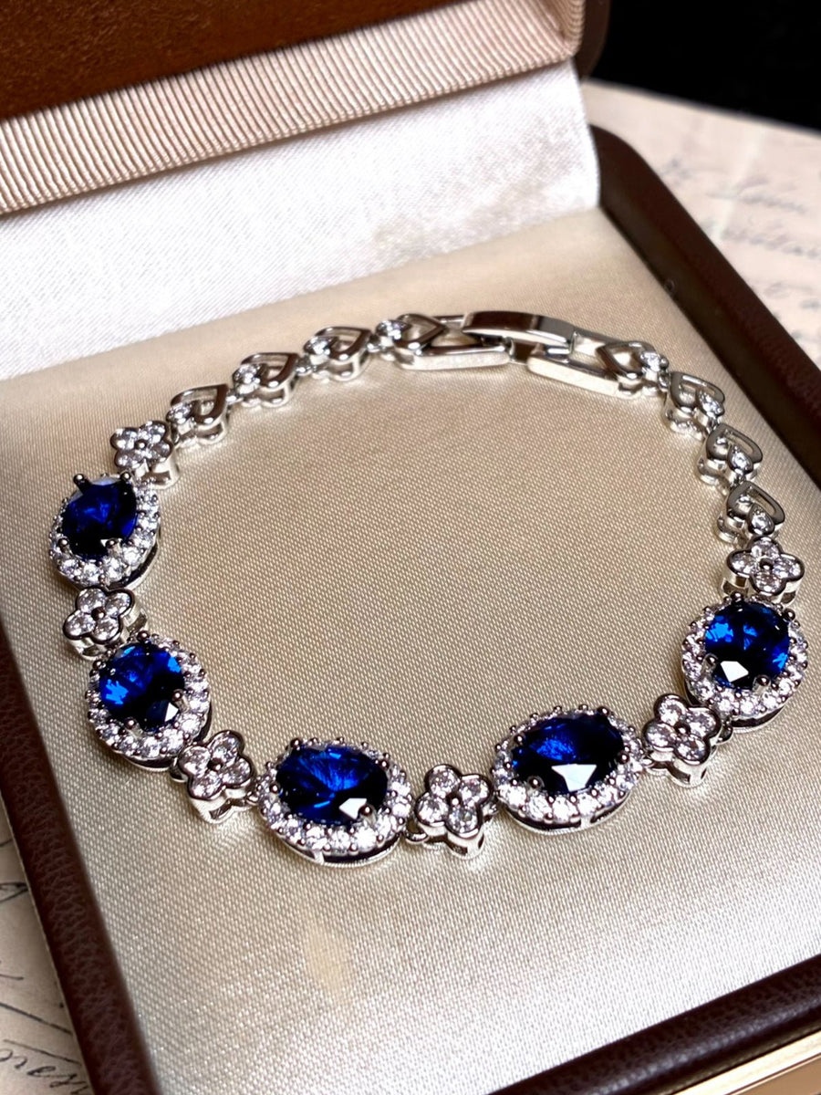 The Social Queen Sapphire Crystal Bracelet features oval sapphire blue crystals in a delicate halo design, plated with white gold and elegantly presented in an open jewelry box on a light fabric base.