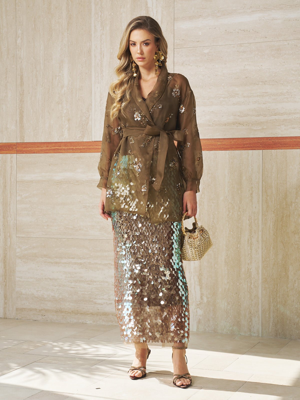 Indoors, a woman wears a sheer brown floral blouse with a waist tie, strappy heels, holds a small woven handbag, and pairs it with the Social Queen Sequin Back-Slit Midi Skirt—perfect for party celebrations.