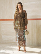 Indoors, a woman wears a sheer brown floral blouse with a waist tie, strappy heels, holds a small woven handbag, and pairs it with the Social Queen Sequin Back-Slit Midi Skirt—perfect for party celebrations.