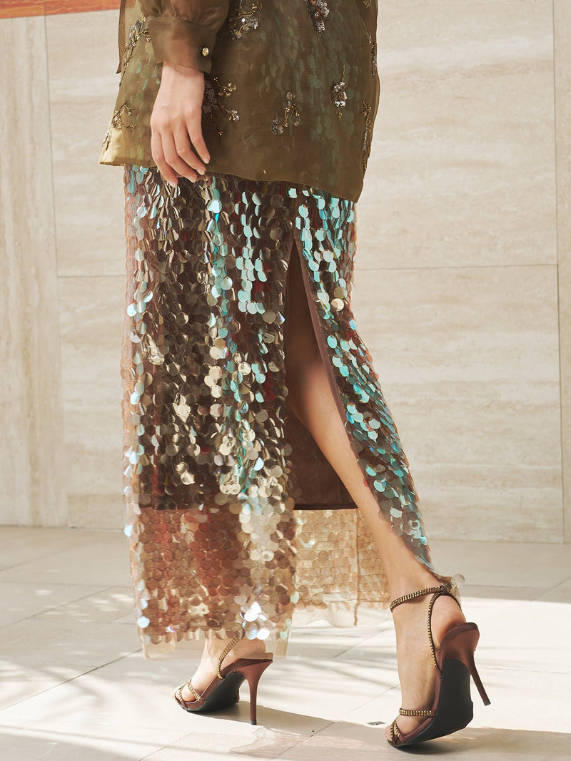 A person wearing a green embellished top, the Social Queen Sequin Back-Slit Midi Skirt, and brown high-heeled sandals stands on a light tile floor.