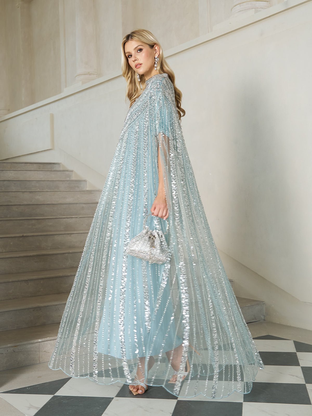 A woman in the Social Queen Sequin Cape Gown with Radiant Beaded Fringe stands on a staircase, accessorized with a small silver handbag and long earrings.