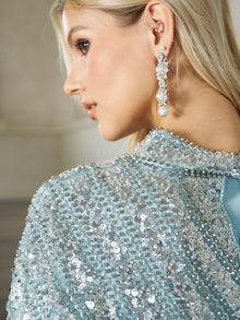 A woman with blonde hair is seen in profile wearing the Social Queen Sequin Cape Gown with Radiant Beaded Fringe, styled with dangling pearl earrings.