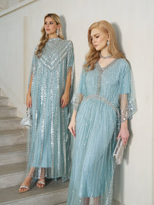 Two women in elegant gowns by a light wall: one, on the stairs with a silver purse, wears Social Queen's Sequin Cape Gown with Radiant Beaded Fringe; the other below carries a silver clutch.