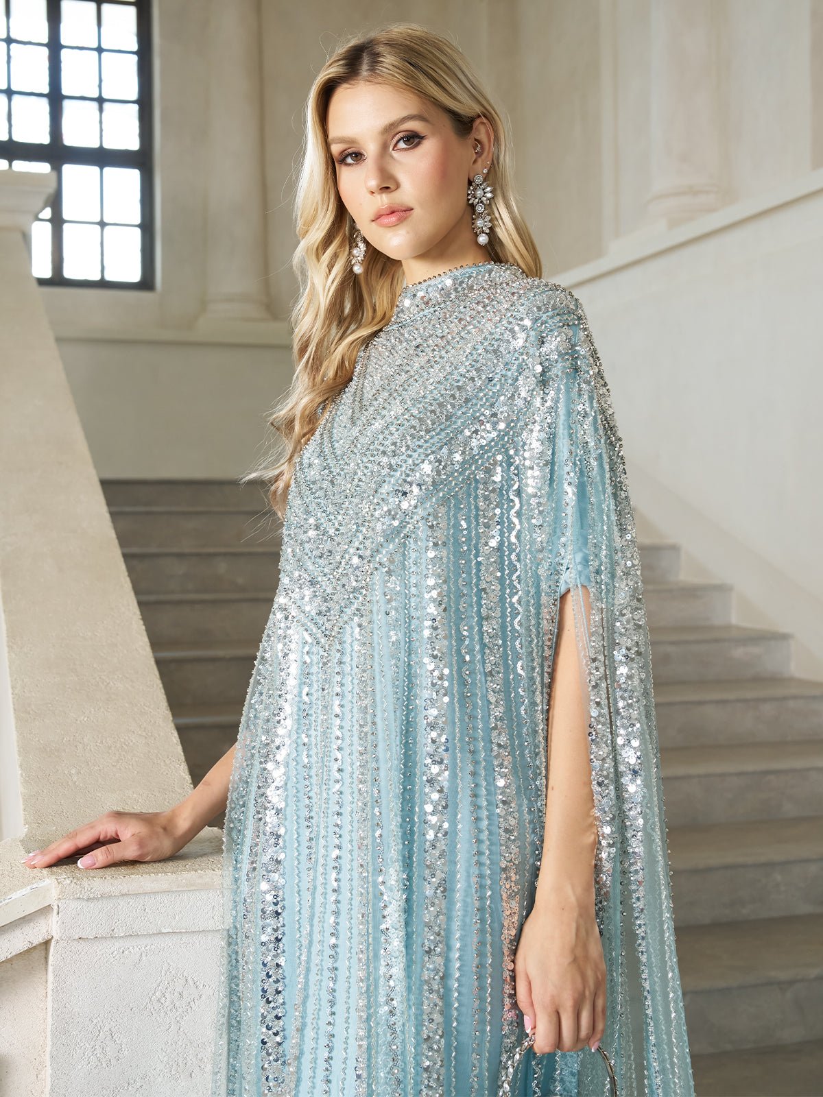 A woman with long blonde hair stands by a staircase in a bright, elegant interior, wearing the Sequin Cape Gown with Radiant Beaded Fringe by Social Queen.