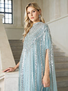 A woman with long blonde hair stands by a staircase in a bright, elegant interior, wearing the Sequin Cape Gown with Radiant Beaded Fringe by Social Queen.
