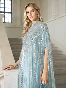 A woman with long blonde hair stands by a staircase in a bright, elegant interior, wearing the Sequin Cape Gown with Radiant Beaded Fringe by Social Queen.