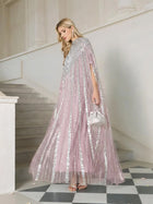 A woman stands on a staircase wearing Social Queen’s Sequin Cape Gown with Radiant Beaded Fringe in pale pink, paired with a silver handbag, silver heels, and drop earrings.