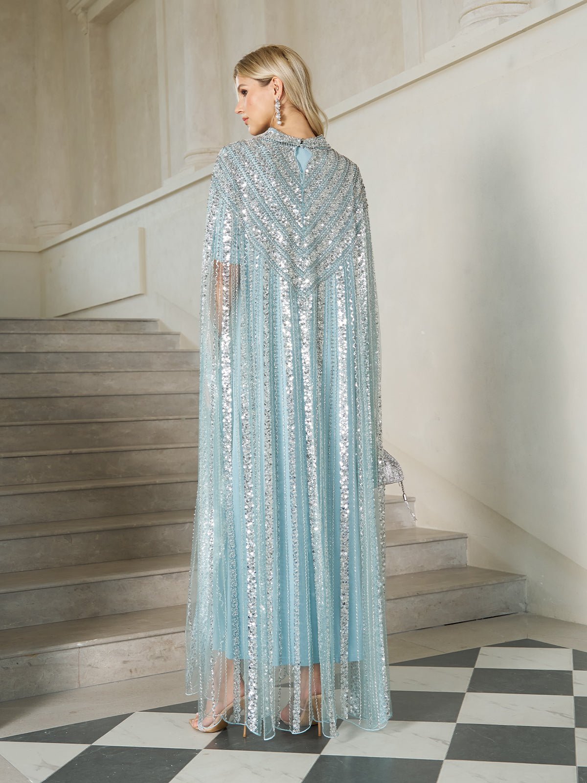 A woman stands on stairs in the Social Queen Sequin Cape Gown with Radiant Beaded Fringe, holding a matching clutch. She faces away, showcasing the elegant back and flowing cape style of this shimmering formal evening dress.
