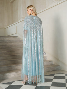 A woman stands on stairs in the Social Queen Sequin Cape Gown with Radiant Beaded Fringe, holding a matching clutch. She faces away, showcasing the elegant back and flowing cape style of this shimmering formal evening dress.