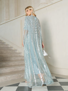 A woman stands on black and white checkered flooring near a staircase, wearing the Social Queen Sequin Cape Gown with Radiant Beaded Fringe and holding a small matching purse.