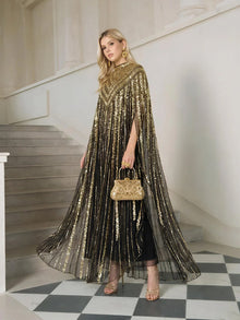 A woman stands on a staircase wearing Social Queen’s Sequin Cape Gown with Radiant Beaded Fringe, paired with a gold handbag, gold heels, and gold earrings.