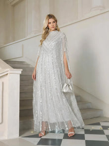 A woman stands on a staircase in the Sequin Cape Gown with Radiant Beaded Fringe by Social Queen, holding a matching small handbag—a stunning choice for any formal occasion.
