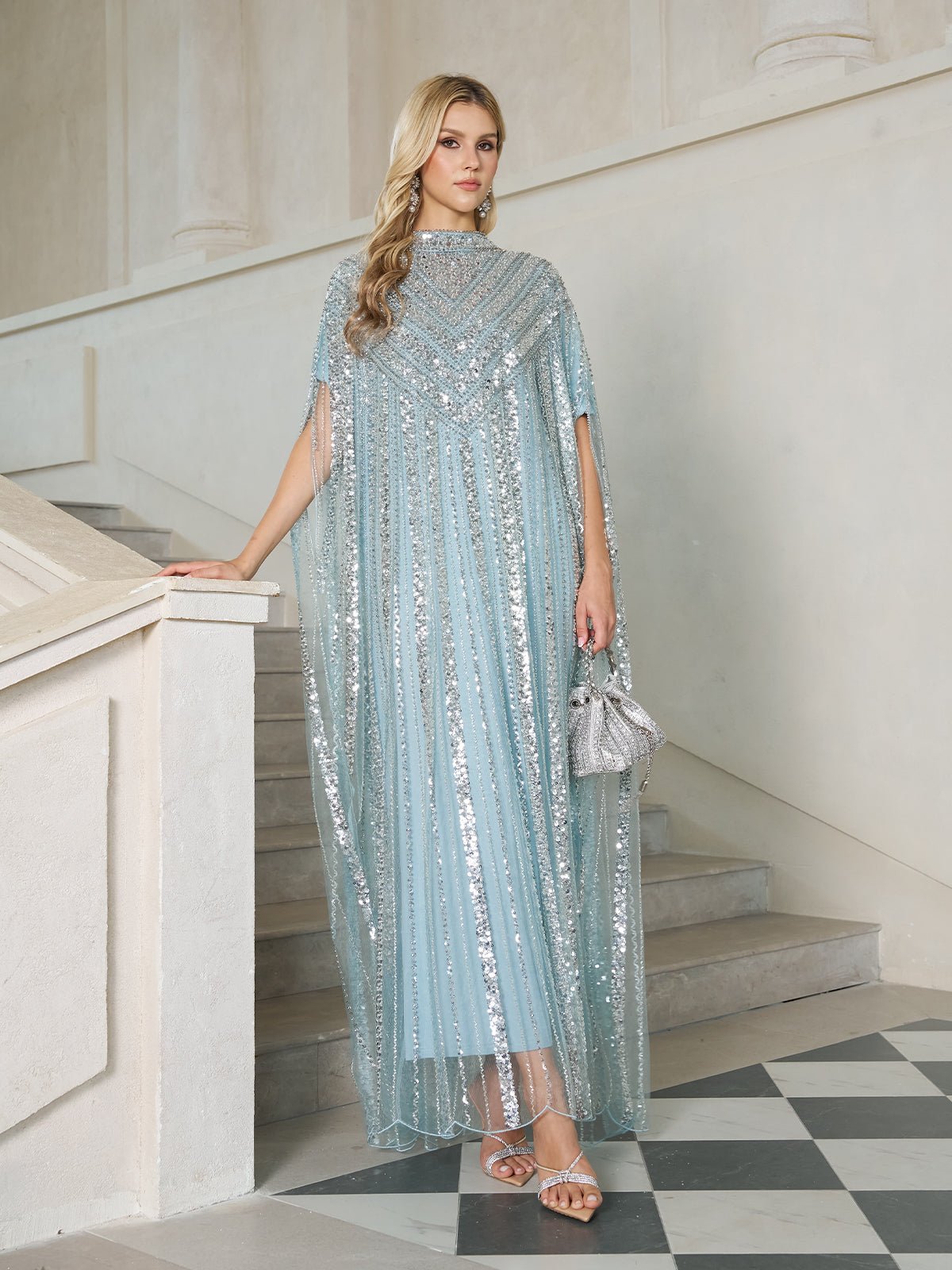 A woman in the Social Queen Sequin Cape Gown with Radiant Beaded Fringe stands on a staircase, holding a silver purse and wearing sandals.