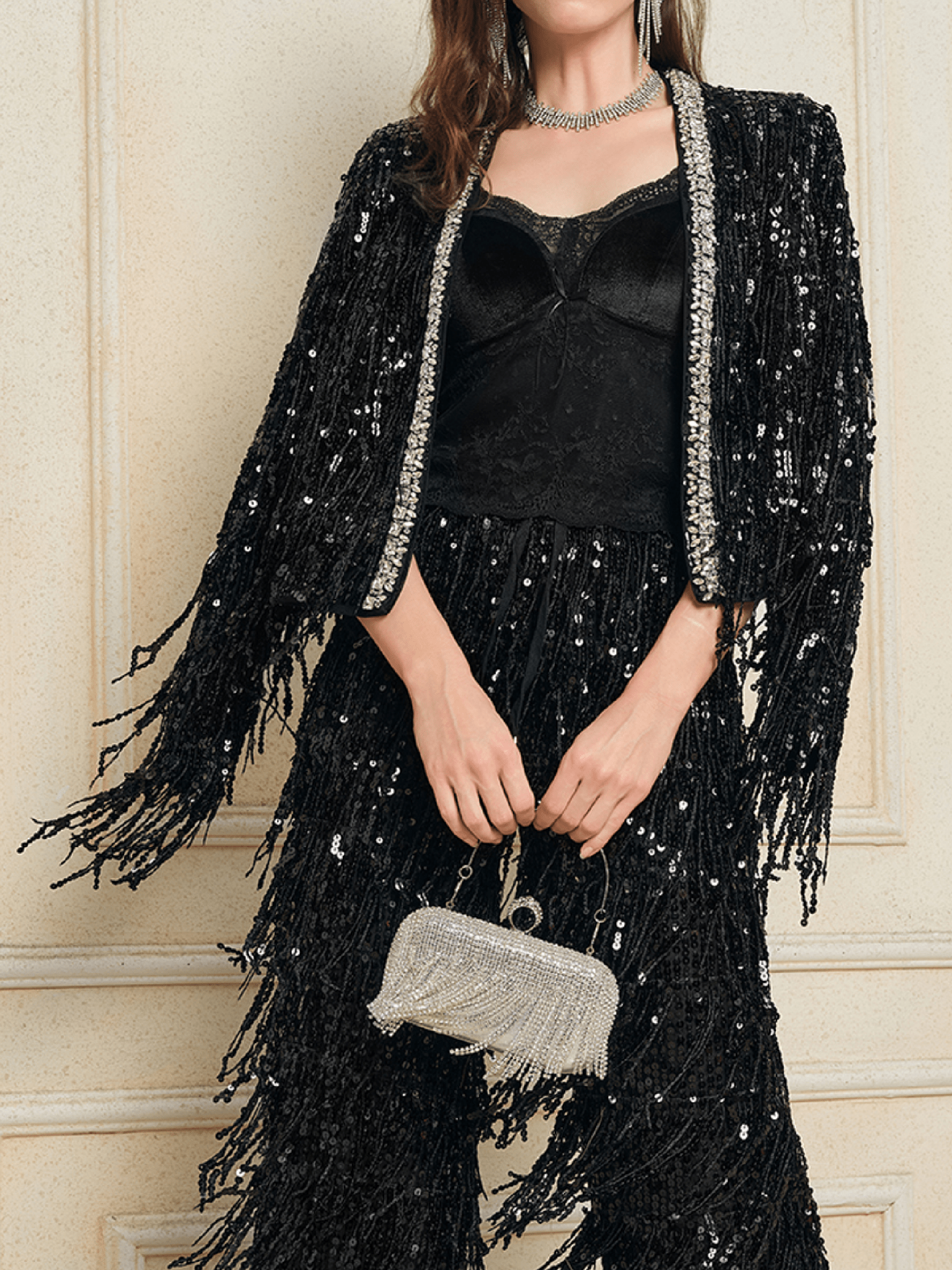 A person wears the Social Queen Sequin & Crystal Fringe Cardigan, paired with a silver necklace and small silver handbag, standing against a cream-colored wall.