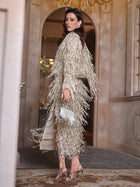 A woman models the Social Queen Sequin & Crystal Fringe Cardigan indoors near a doorway, accessorizing with a small silver purse and strappy high-heeled sandals.