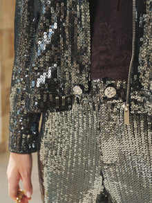 Close-up of a person wearing the Sequin-Embellished Metallic Crop Jacket by Social Queen, partially unzipped, paired with matching pants; hand resting at their side.