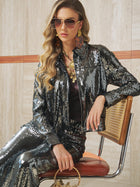A woman sits on a chair in a Social Queen Sequin-Embellished Metallic Crop Jacket, paired with matching pants, aviator sunglasses, gold jewelry, and holding a small handbag.