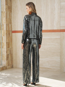 A woman stands indoors with her back to the camera, wearing Social Queen's Sequin-Embellished Wide-Leg Pants, paired with a shiny black sequin jacket, black heels, and large earrings.