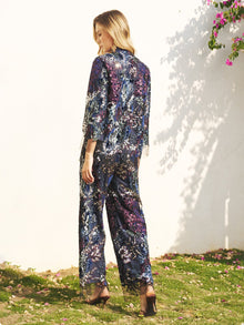 A woman stands on grass facing away, wearing Social Queen’s Sequin Embroidered Mesh Wide-Leg Pants in dark blue. Sunlight throws her shadow onto a white wall as she shows off the pants' sheer, floral-patterned fabric outdoors.