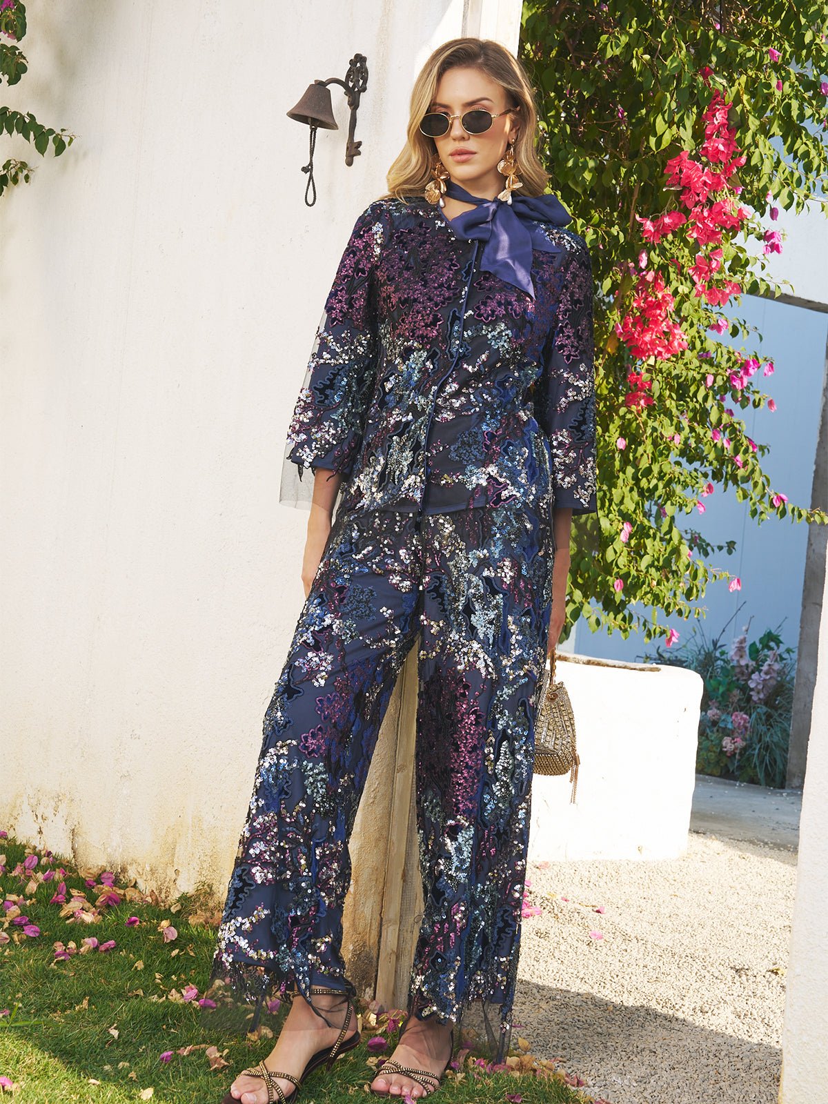 A woman stands outdoors wearing Social Queen’s Sequin Embroidered Mesh Wide-Leg Pants and sunglasses, holding a small handbag amid white walls and pink flowers.