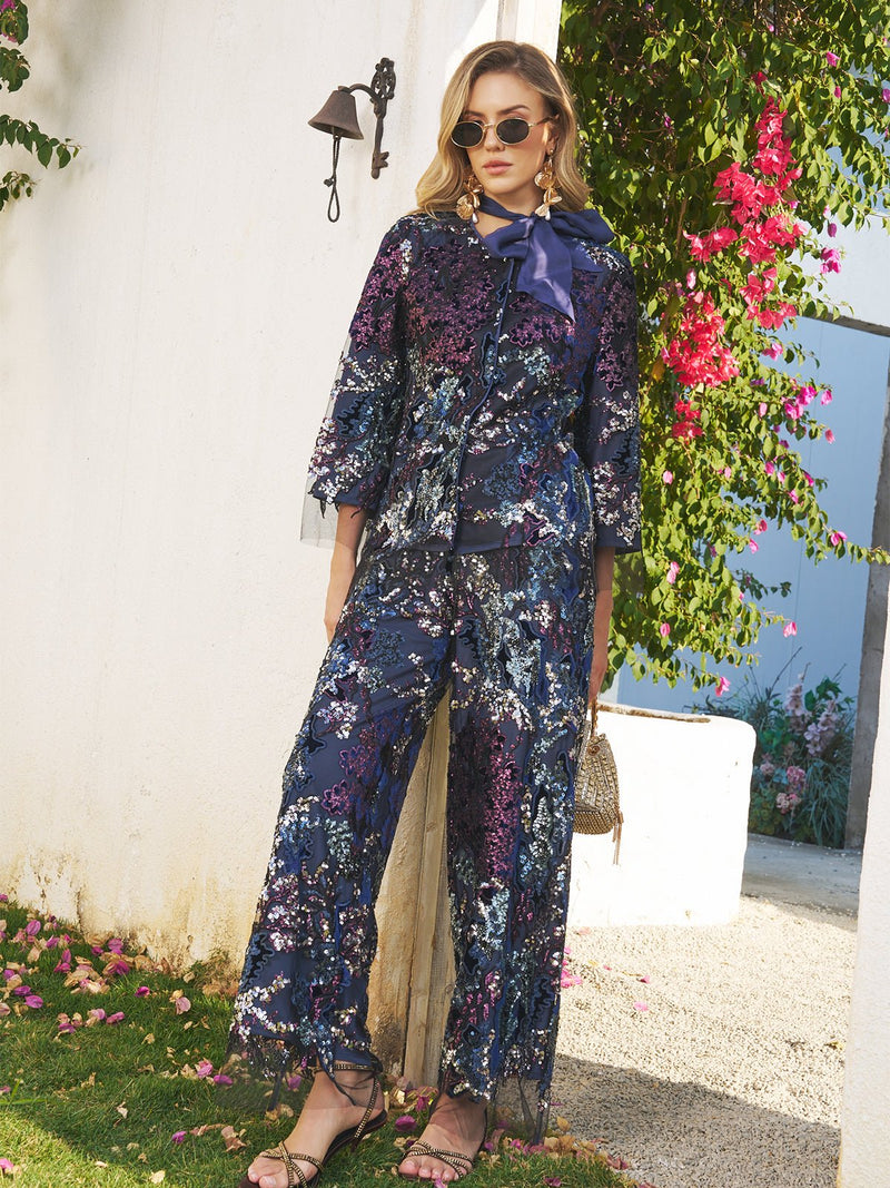 A woman stands outdoors wearing Social Queen’s Sequin Embroidered Mesh Wide-Leg Pants and sunglasses, holding a small handbag amid white walls and pink flowers.