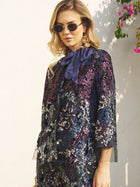A woman in sunglasses and gold earrings wears the Social Queen Sequin Embroidered Velvet Mesh Jacket with a navy bow, posing against a light wall with pink flowers—perfect for party celebrations.
