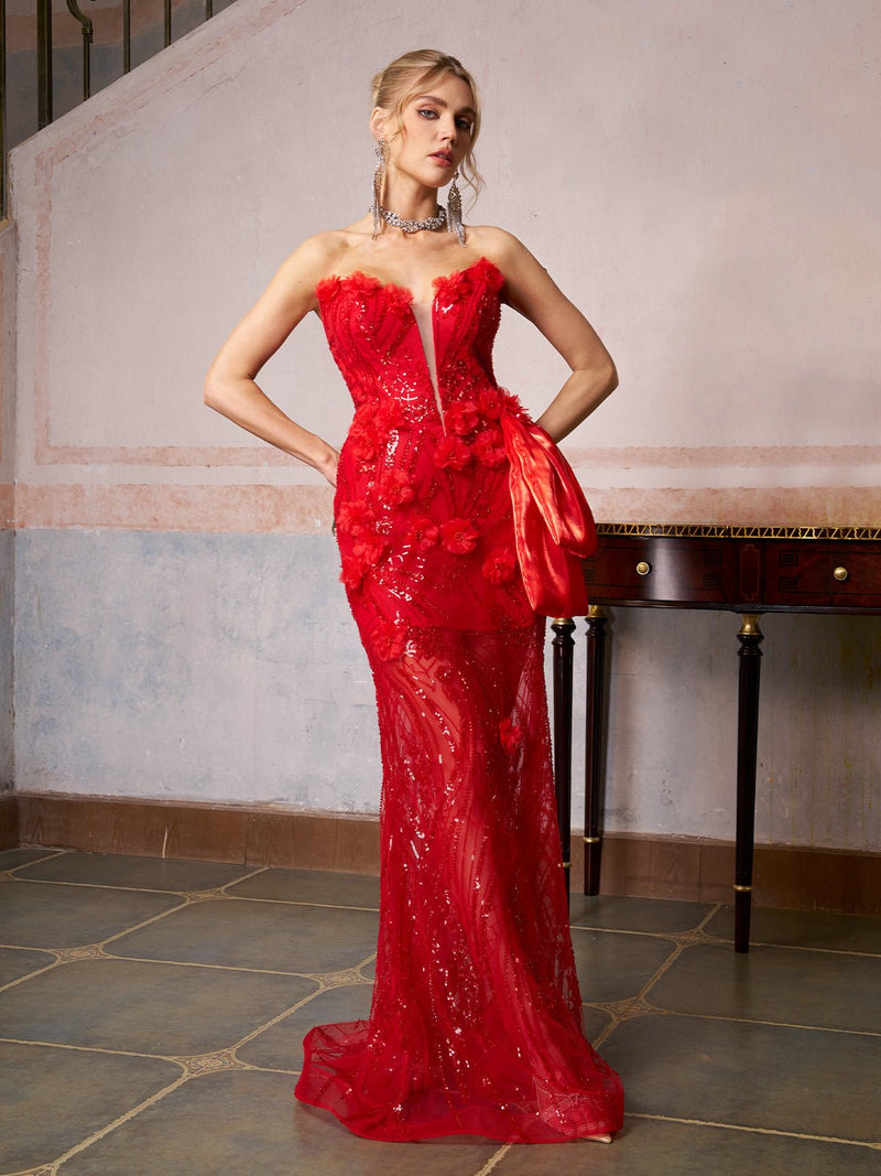 A woman wears the Sequin Floral Mermaid Gown by Social Queen, featuring a strapless fit, floral appliqués, sequins, and a side bow, as she poses indoors with hands on hips in front of a console table and painted wall.