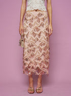 A woman stands against a pink background wearing the Social Queen Sequin Fringe Mesh Skirt with a cream lace top, holding a small gold purse and wearing strappy sandals.