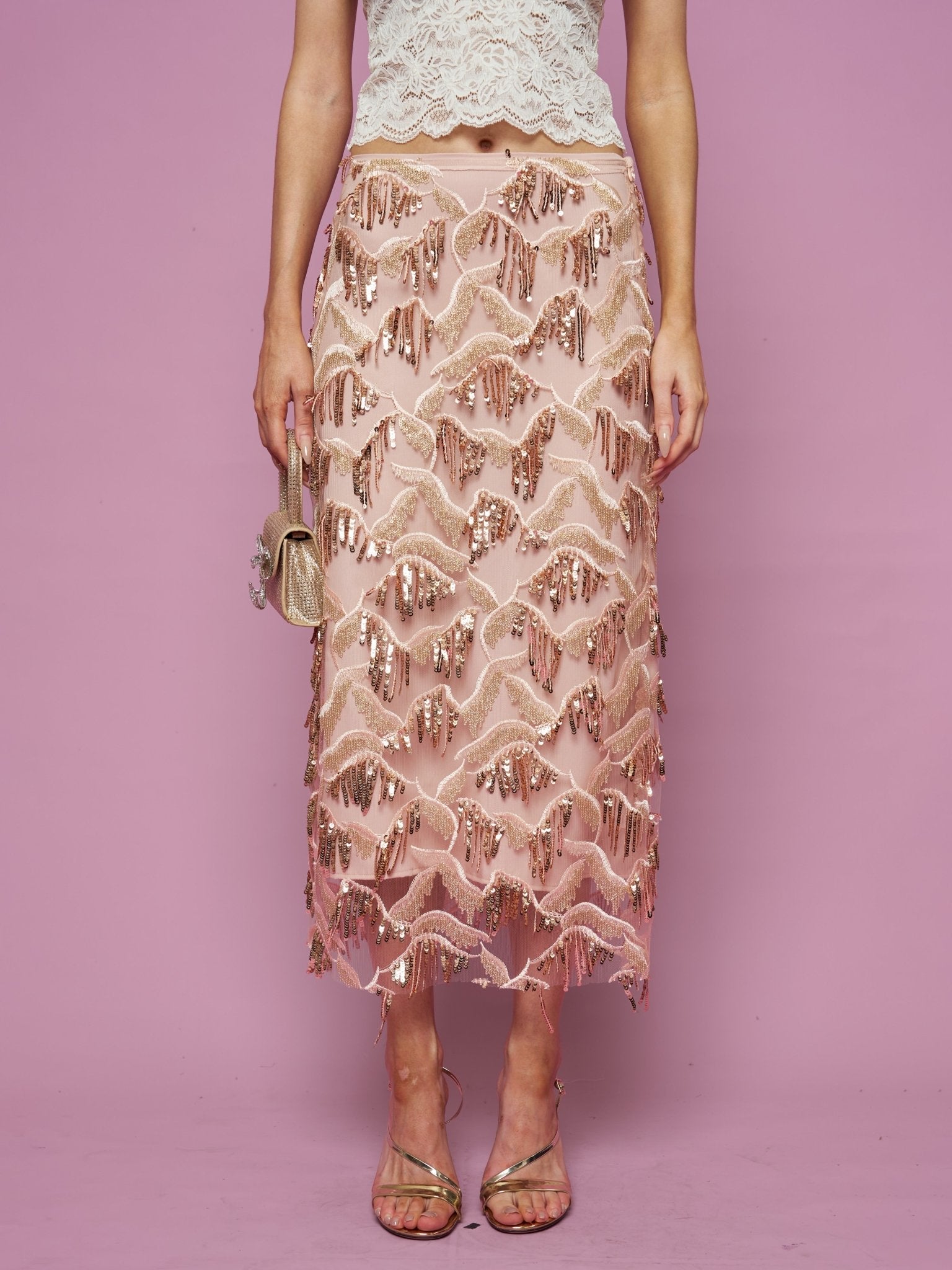 A woman stands against a pink background wearing the Social Queen Sequin Fringe Mesh Skirt with a cream lace top, holding a small gold purse and wearing strappy sandals.