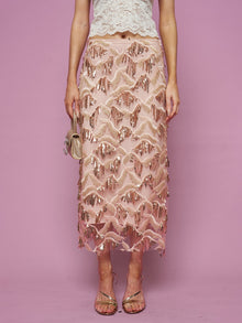 A woman stands against a pink background wearing the Social Queen Sequin Fringe Mesh Skirt with a cream lace top, holding a small gold purse and wearing strappy sandals.