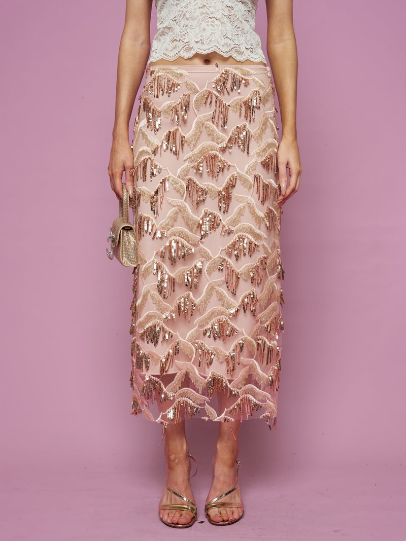 A woman stands against a pink background wearing the Social Queen Sequin Fringe Mesh Skirt with a cream lace top, holding a small gold purse and wearing strappy sandals.