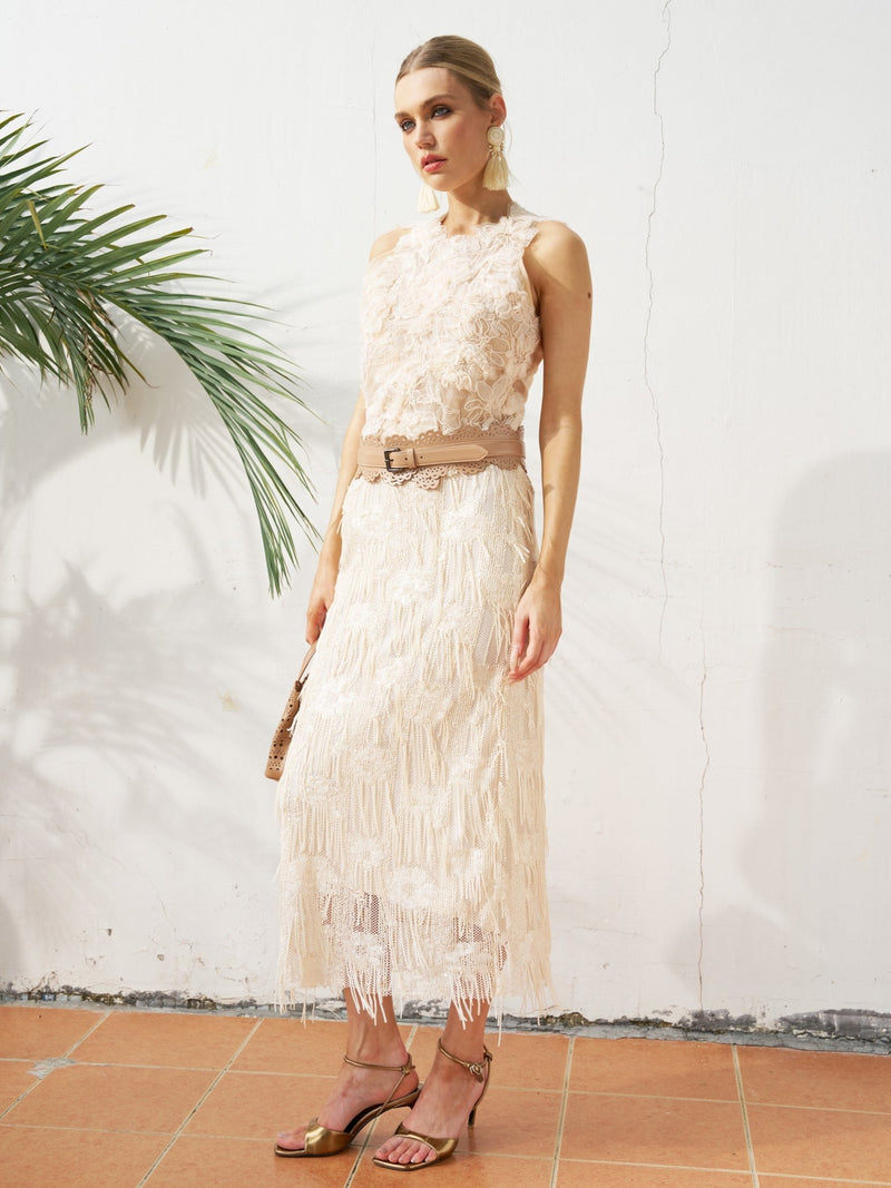A woman stands indoors wearing the Social Queen Sequin Fringe Mesh Skirt with a belt, holding a small bag and heeled sandals. A palm leaf on the left highlights effortless spring-summer styling.