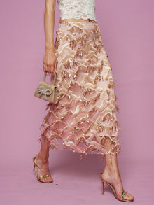 A person stands against a pink background, wearing a white lace top, Social Queen Sequin Fringe Mesh Skirt, gold high-heeled sandals, and holding a small sparkly handbag.