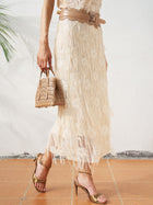 A woman in the Social Queen Sequin Fringe Mesh Skirt and gold heels, perfect for spring-summer looks, holds a small tan bag while standing on a tiled floor.