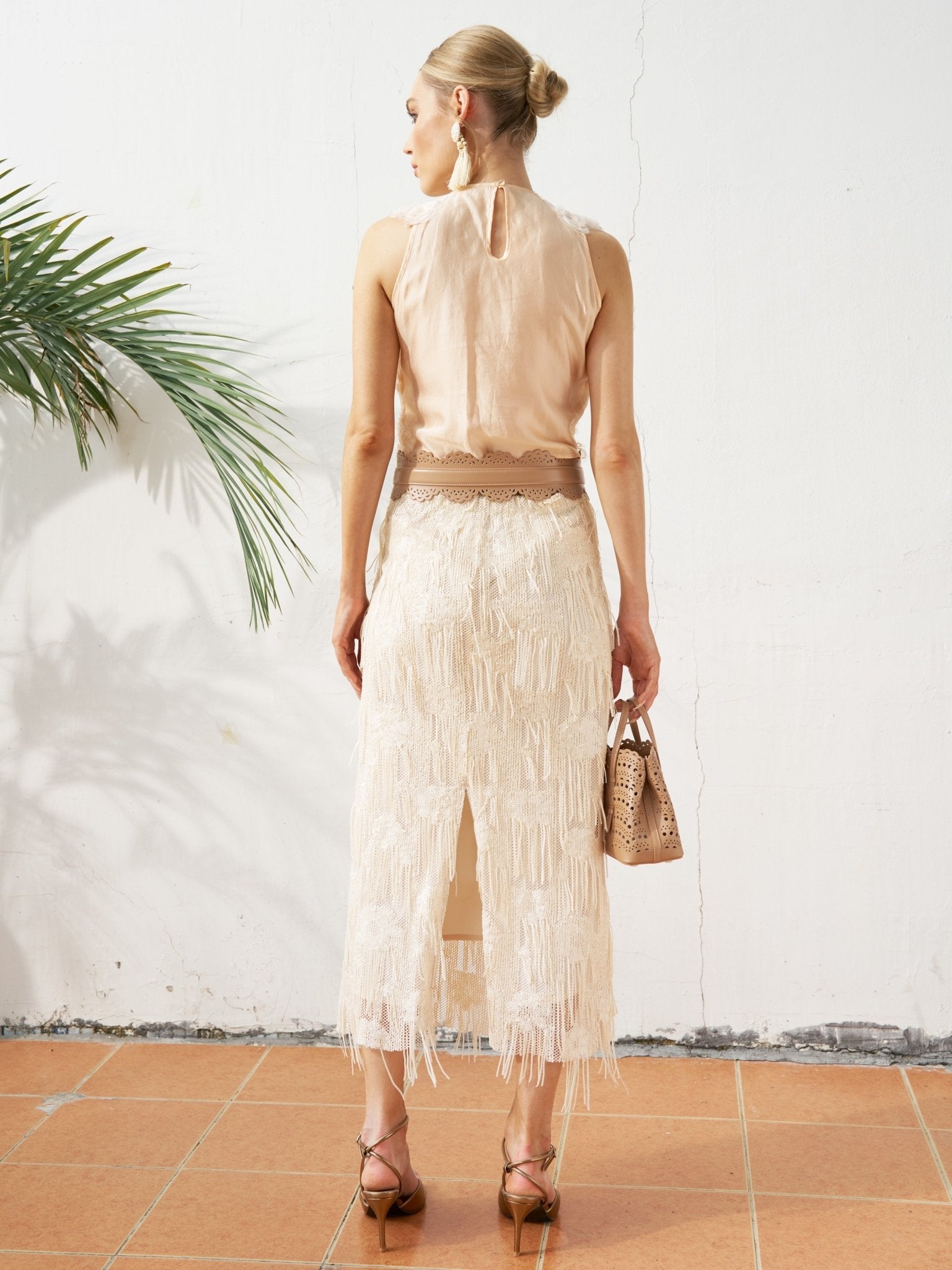 A woman stands indoors facing away, styled for spring/summer in a sleeveless beige top and the Sequin Fringe Mesh Skirt by Social Queen, holding a small handbag with a potted plant to her left.