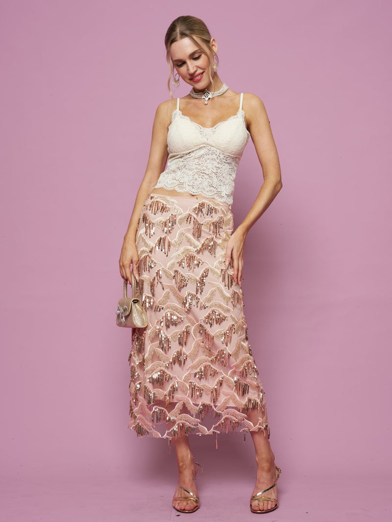 Woman poses against a pink background wearing a white lace top, the Social Queen Sequin Fringe Mesh Skirt in pink with a high-rise straight silhouette, heeled sandals, and holding a small purse.
