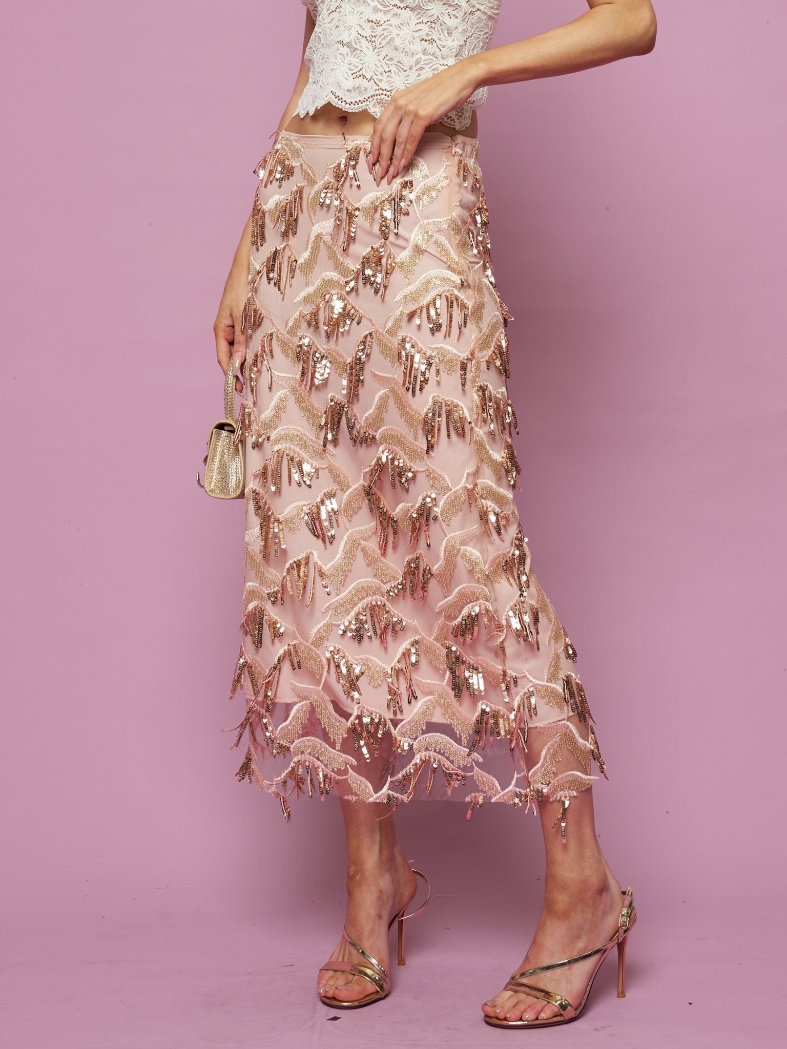 A person in a white lace crop top and the Social Queen Sequin Fringe Mesh Skirt stands against a pink background, holding a small gold purse.