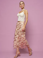 A woman in a white lace top and the Social Queen Sequin Fringe Mesh Skirt stands against a pink background, accessorized with gold high-heeled sandals and jewelry.