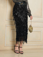 A person in the Social Queen Sequin Fringe Side-Slit Midi Skirt, black strappy heels, a metallic long-sleeve top, and holding a small gold purse stands against a beige wall.