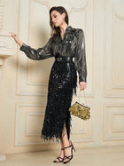 A woman stands indoors against a cream wall, wearing Social Queen’s Sequin Fringe Side-Slit Midi Skirt with a shiny metallic blouse, black belt, strappy heels, and holding a gold embellished handbag.