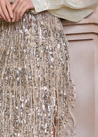 A close-up shows a hand resting on the Social Queen Sequin Fringe Side-Slit Midi Skirt, featuring fringe trim and pearl buttons, against an ornate, light-toned background.