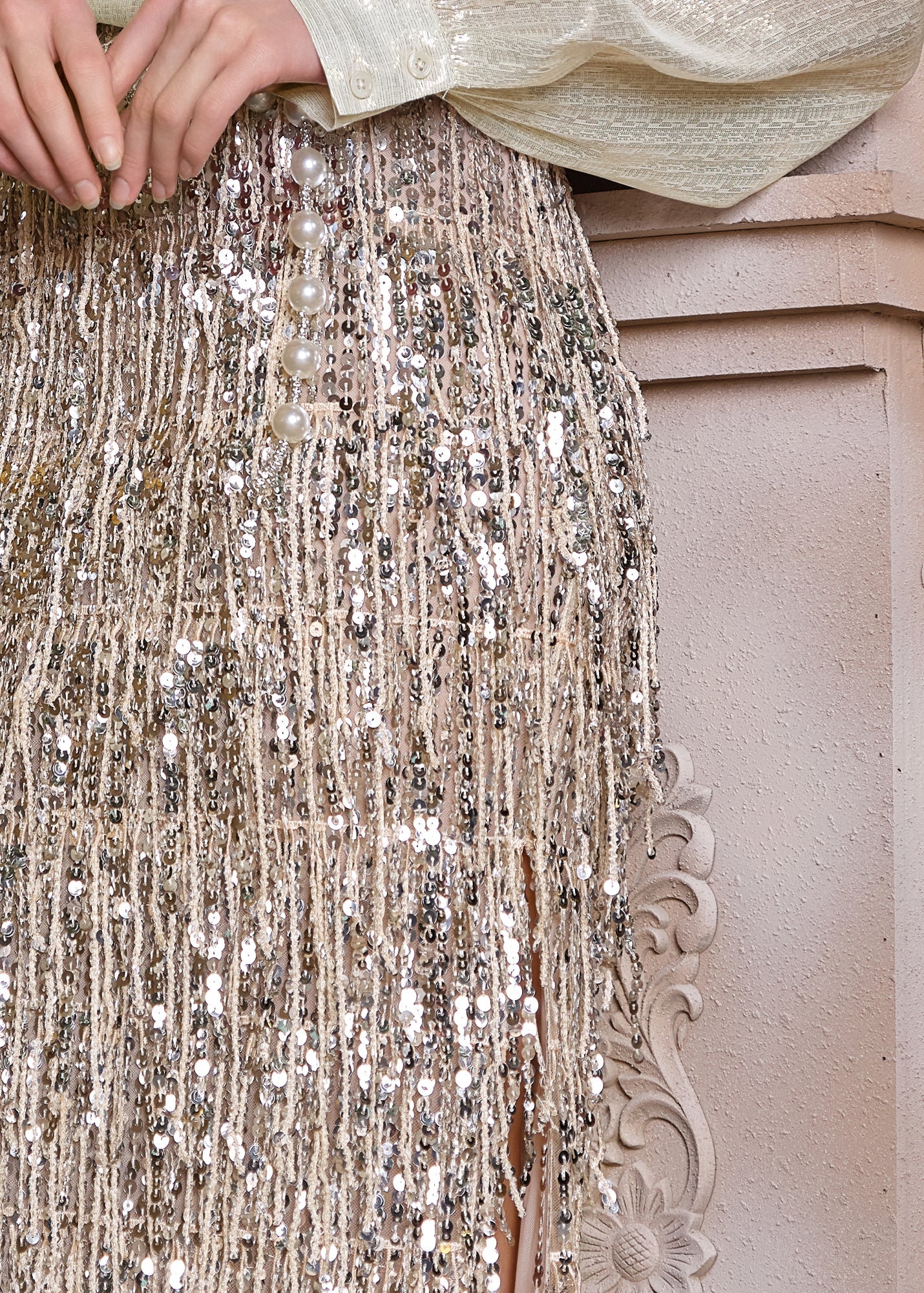 A close-up shows a hand resting on the Social Queen Sequin Fringe Side-Slit Midi Skirt, featuring fringe trim and pearl buttons, against an ornate, light-toned background.